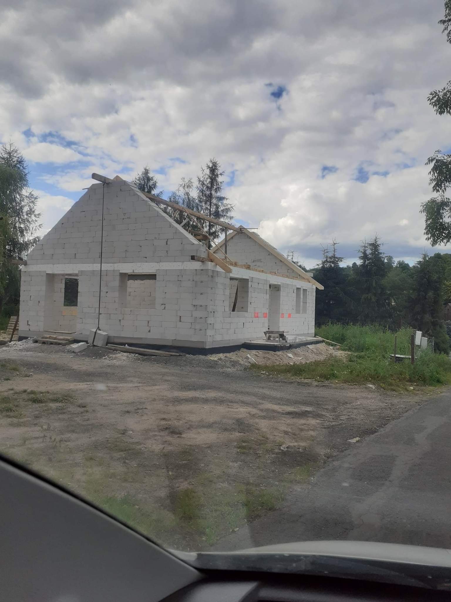 Surowy stan budynku w trakcie budowy, ściany wzniesione z białych bloczków, widoczne otwory okienne i drzwiowe, częściowo wykonana konstrukcja dachu, tło stanowią drzewa i pochmurne niebo.