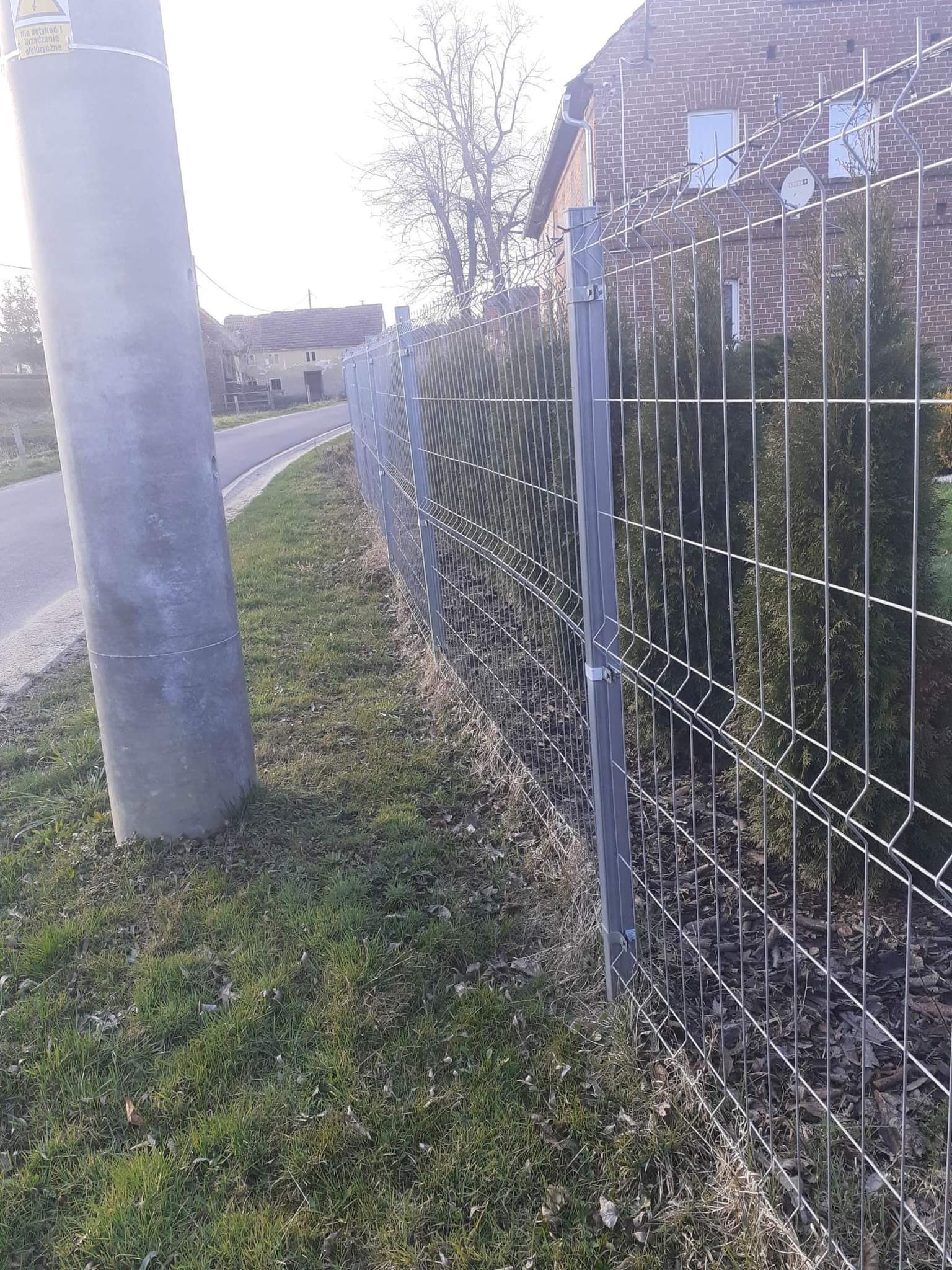 Szara, metalowa siatka ogrodzeniowa panelowa zamontowana wzdłuż drogi, z widocznymi słupkami i elementami mocującymi, za ogrodzeniem posadzone tuje, w tle budynek z czerwonej cegły i słup...