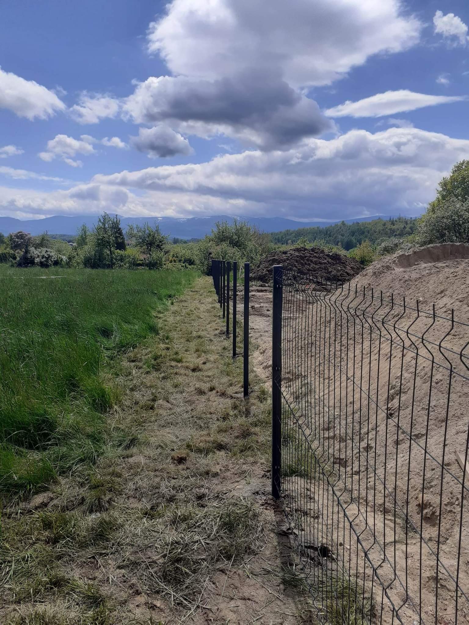 Montaż czarnego ogrodzenia panelowego na granicy działki z wysoką trawą i usypanym wzgórzem ziemi w tle, widok na góry pod błękitnym, pochmurnym niebem.