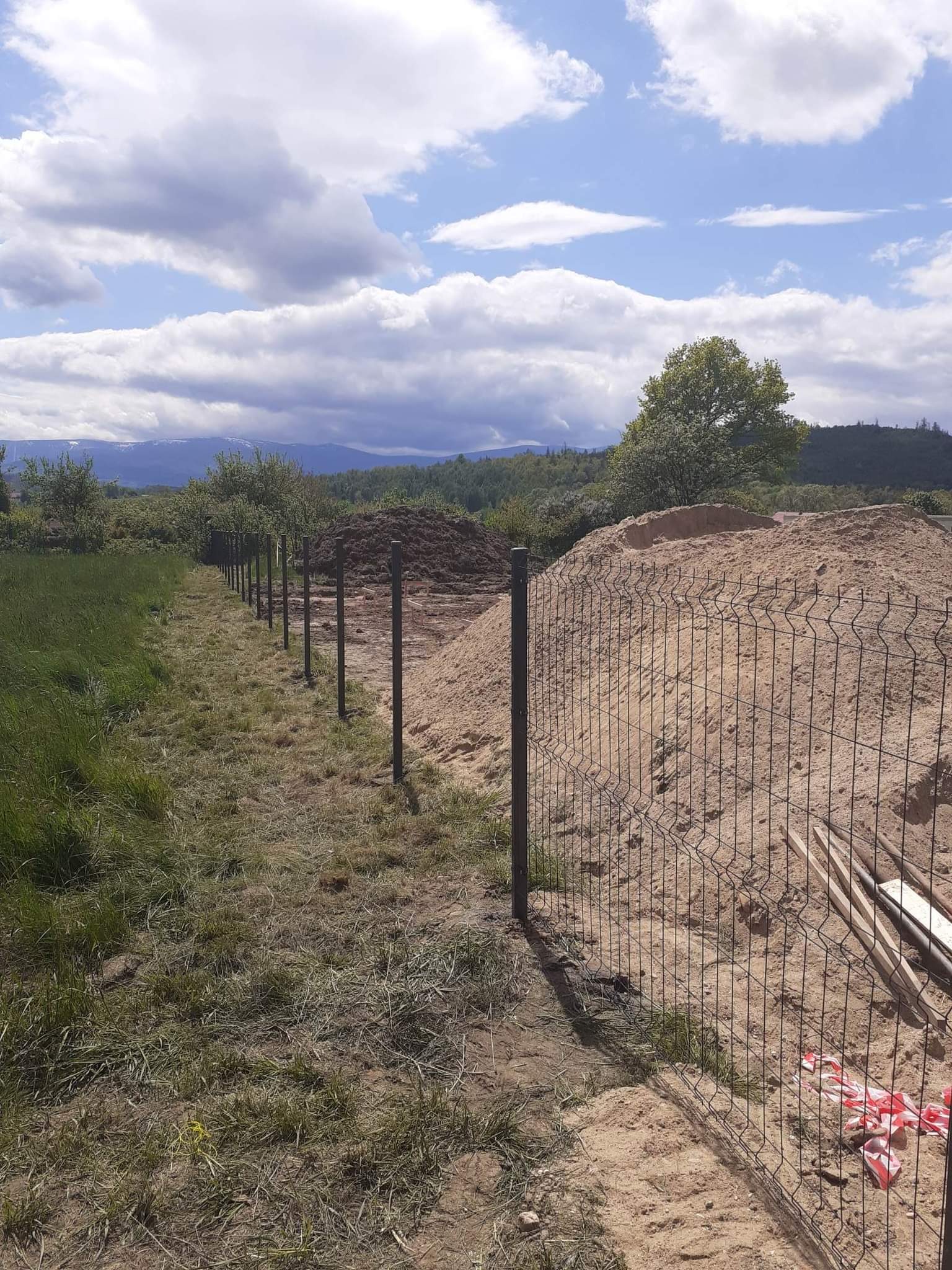 Montaż metalowej siatki ogrodzeniowej na słupkach w terenie z przygotowanymi nasypami ziemi, widok na odległe góry i pochmurne niebo.