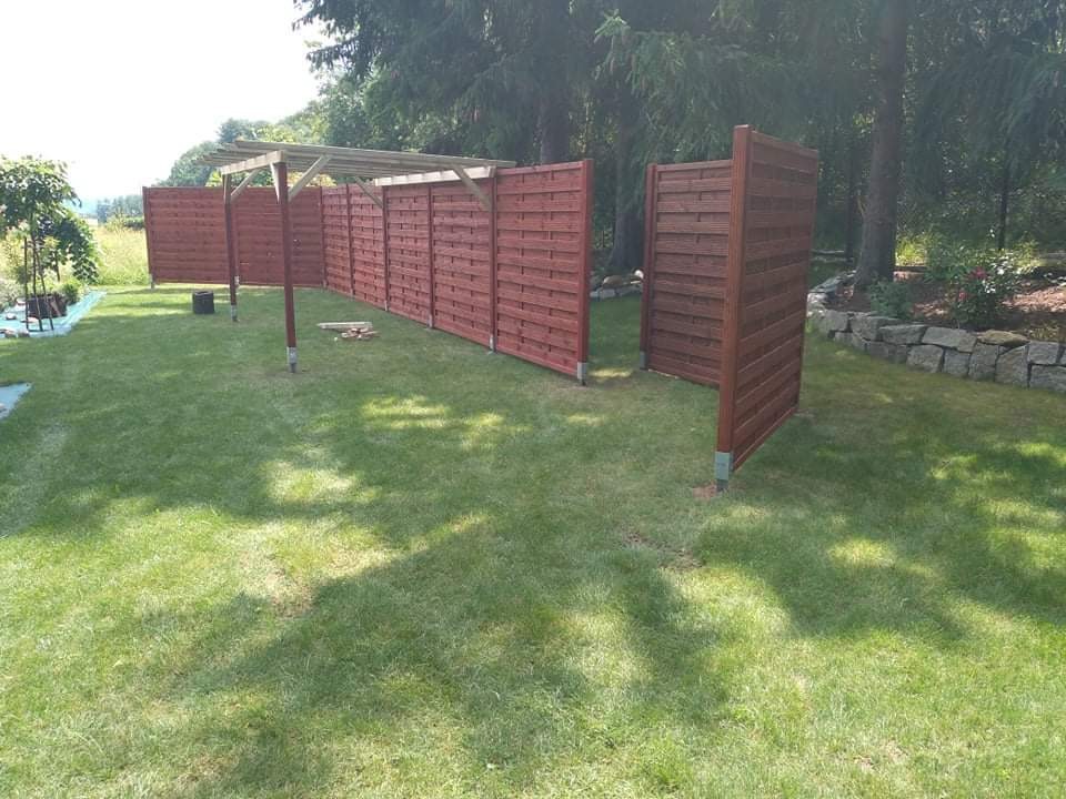 Brązowe, drewniane panele ogrodzeniowe z metalowymi słupkami, częściowo połączone drewnianą konstrukcją pergoli, rozstawione na trawniku w ogrodzie.