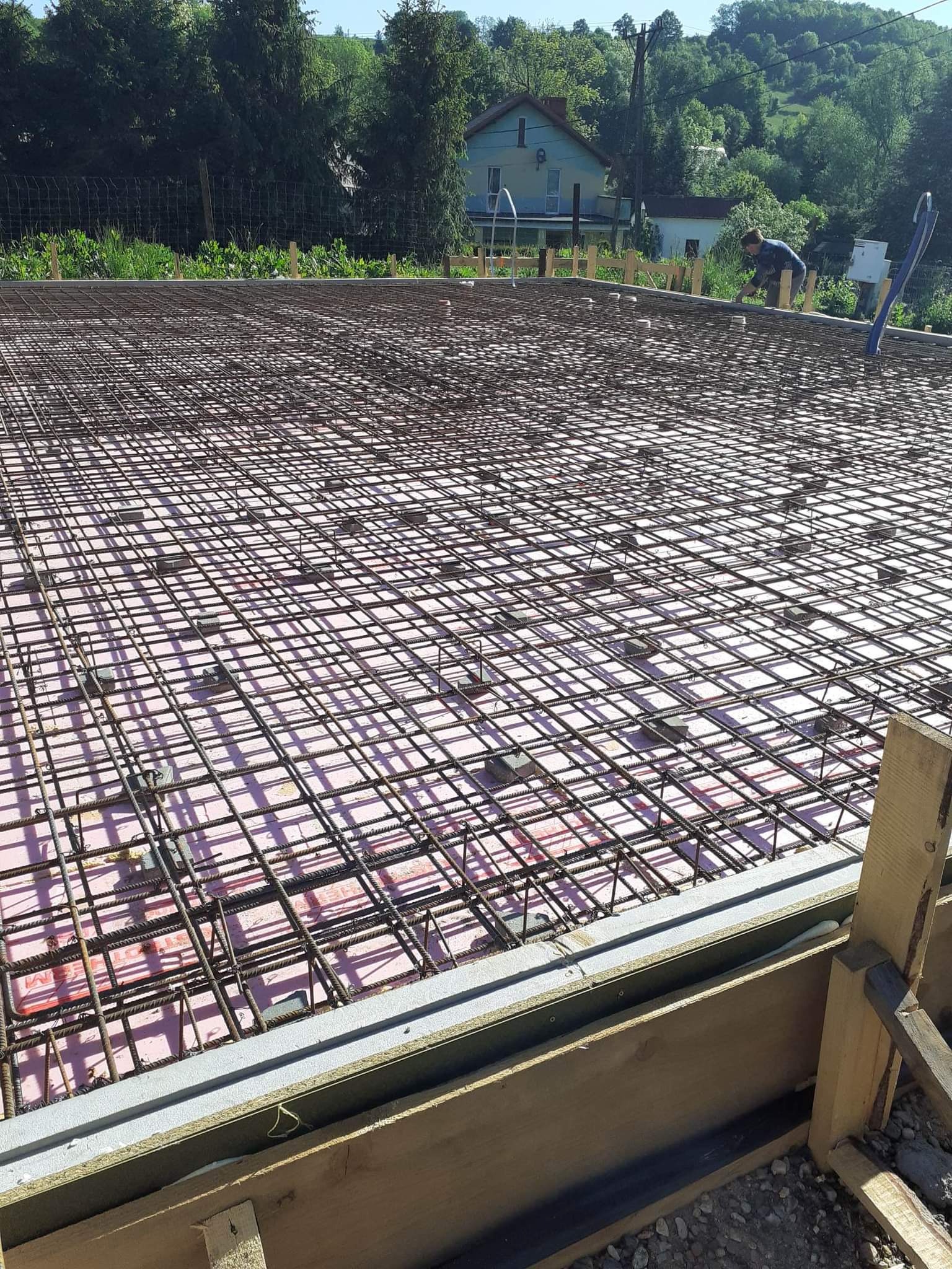 Zbrojenie płyty fundamentowej z siatki stalowej na izolacji termicznej, widoczne szalunki drewniane i pracownik budowlany w tle.