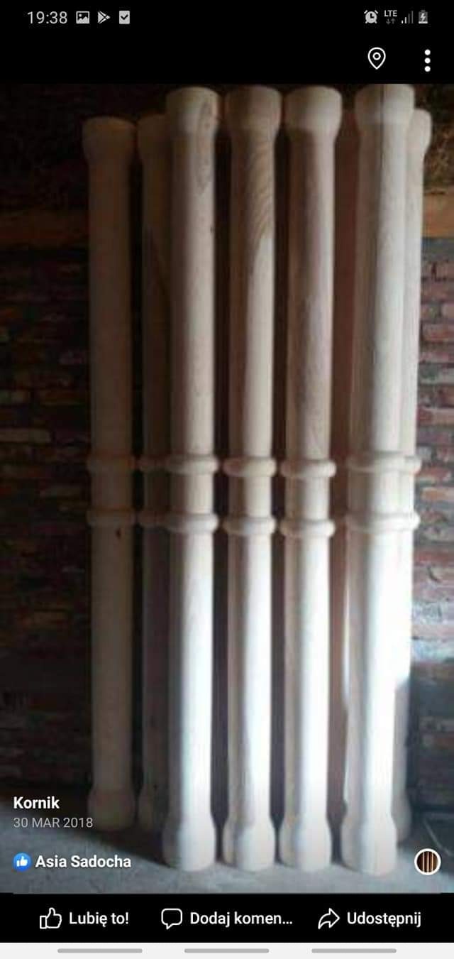 Surowe, nielakierowane drewniane kolumny z dekoracyjnymi elementami, ustawione pionowo w rzędach przed ceglaną ścianą.