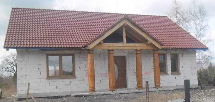 Surowy stan domu z czerwonym dachem i drewnianym gankiem z kolumnami, widoczne okna z brązowymi ramami i drzwi wejściowe, otoczenie w stanie naturalnym.