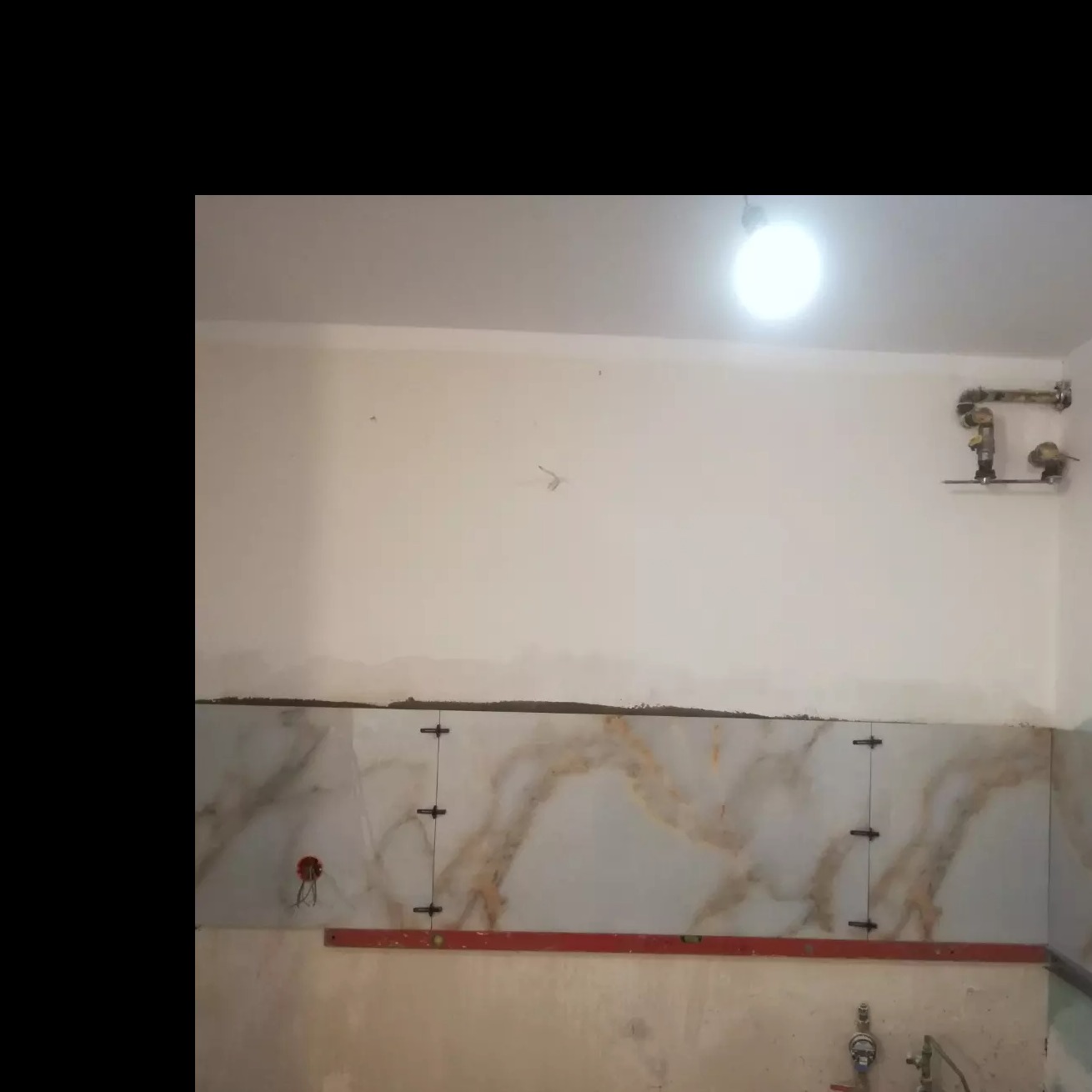 Fragment ściany z częściowo ułożonymi płytkami imitującymi marmur, poziomica i krzyżyki dystansowe w trakcie prac wykończeniowych w pomieszczeniu.