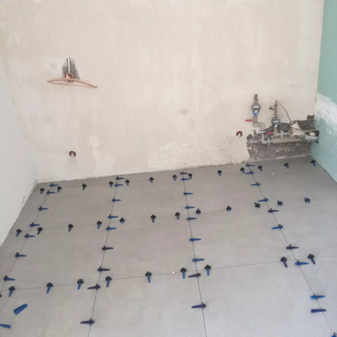 Przygotowanie podłogi do ułożenia płytek z użyciem systemu poziomowania, widoczne nieotynkowane ściany i wystające rury.