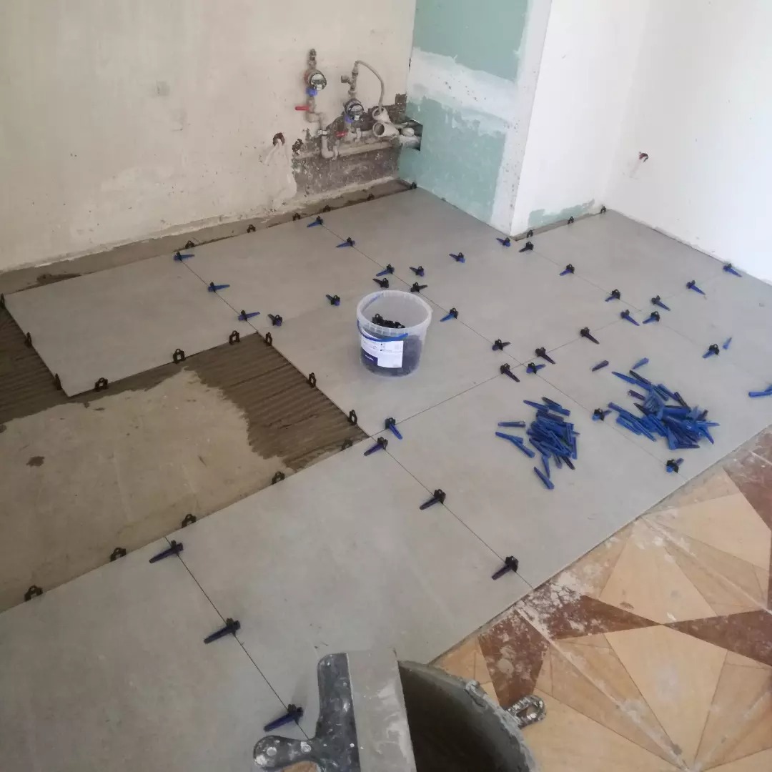 Proces układania szarych płytek podłogowych w pomieszczeniu, widoczne krzyżyki dystansowe, wiadro z zaprawą i fragment starej podłogi z wzorem geometrycznym.