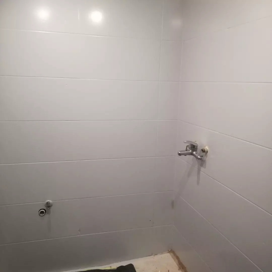 Wnętrze łazienki w trakcie wykańczania, widoczne białe, poziomo ułożone płytki na ścianach, przygotowane podejścia wodne i kanalizacyjne, surowy stan podłogi.