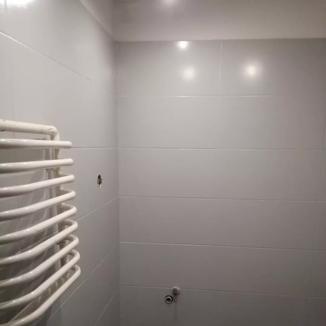 Biała łazienka z poziomymi płytkami, widoczny biały grzejnik drabinkowy i otwór w ścianie po instalacji.