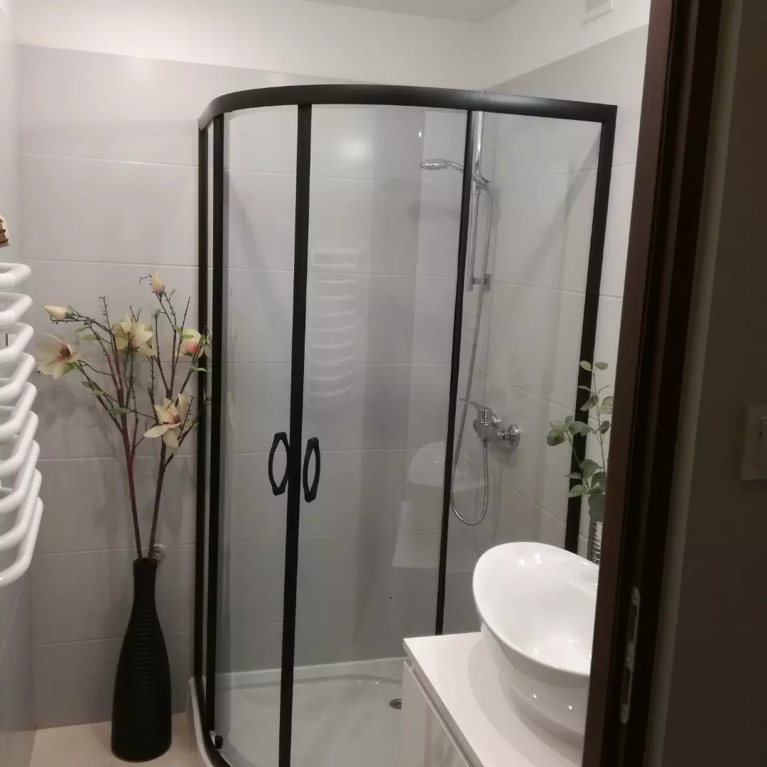 Nowoczesna kabina prysznicowa z czarną ramą, białe płytki na ścianach, dekoracyjny wazon z kwiatami i umywalka nablatowa w wykończonej łazience.