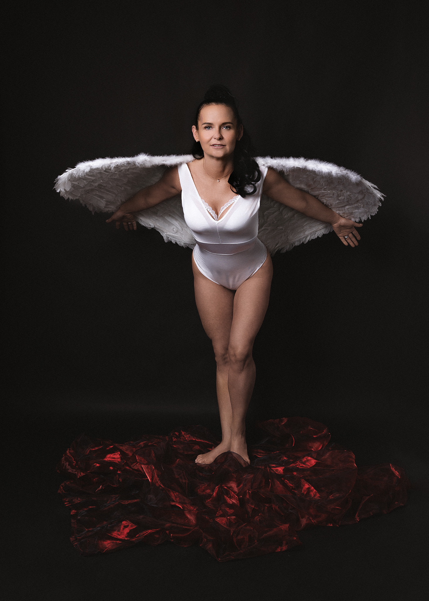 Kobieta w białym body i skrzydłach anioła, stojąca na czerwonej, błyszczącej tkaninie na czarnym tle, uśmiechnięta, z włosami upiętymi do góry.