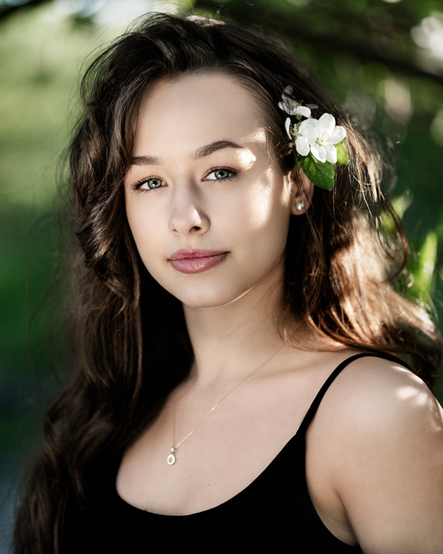 Portret młodej kobiety z kwiatem jabłoni we włosach, delikatny makijaż, złoty naszyjnik z okrągłą zawieszką na tle zieleni.