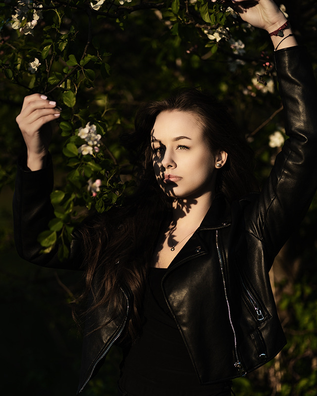 Portret młodej kobiety w czarnej skórzanej kurtce, pozującej w plenerze na tle kwitnącego drzewa, z twarzą częściowo zacienioną przez liście.