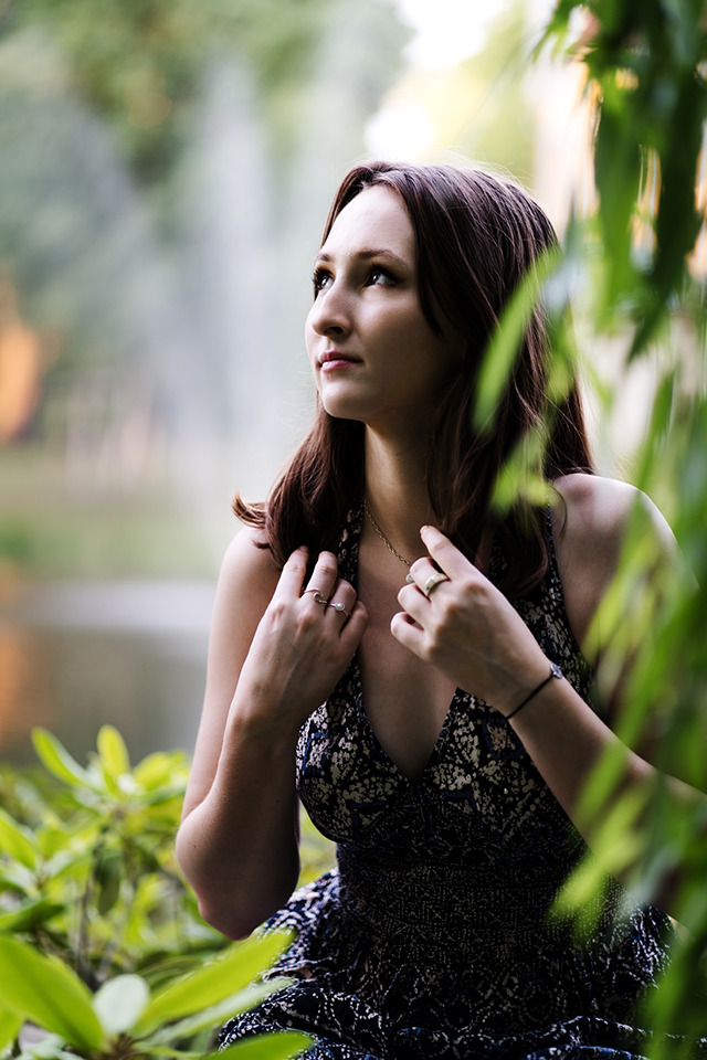 Portret kobiety w kwiecistej sukience, spoglądającej w górę w plenerze, z fontanną w tle i zielonymi liśćmi w pierwszym planie.