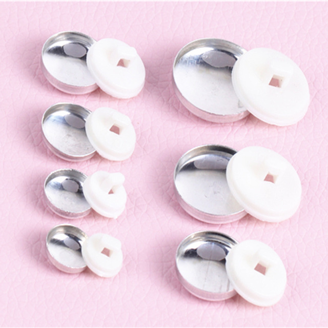 Metalowe zatrzaski z białymi plastikowymi nakładkami na różowym tle, różne rozmiary.