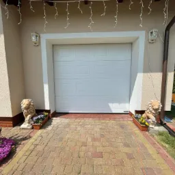 Biała segmentowa konstrukcja z dekoracyjnymi lampionami i kamiennymi lwami po bokach, zdobiąca wjazd do garażu wyłożony kostką brukową w odcieniach beżu i czerwieni.