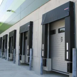 Szereg szarych bram przemysłowych z czarnymi osłonami i numerem '6' na jednej z nich, widoczne z zewnątrz hali magazynowej.