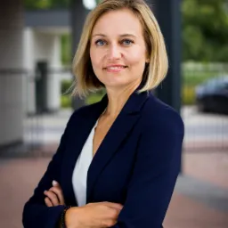 Portret uśmiechniętej kobiety w granatowej marynarce, z założonymi rękami i bransoletką z ciemnych koralików, na tle nowoczesnego budynku z zielenią.