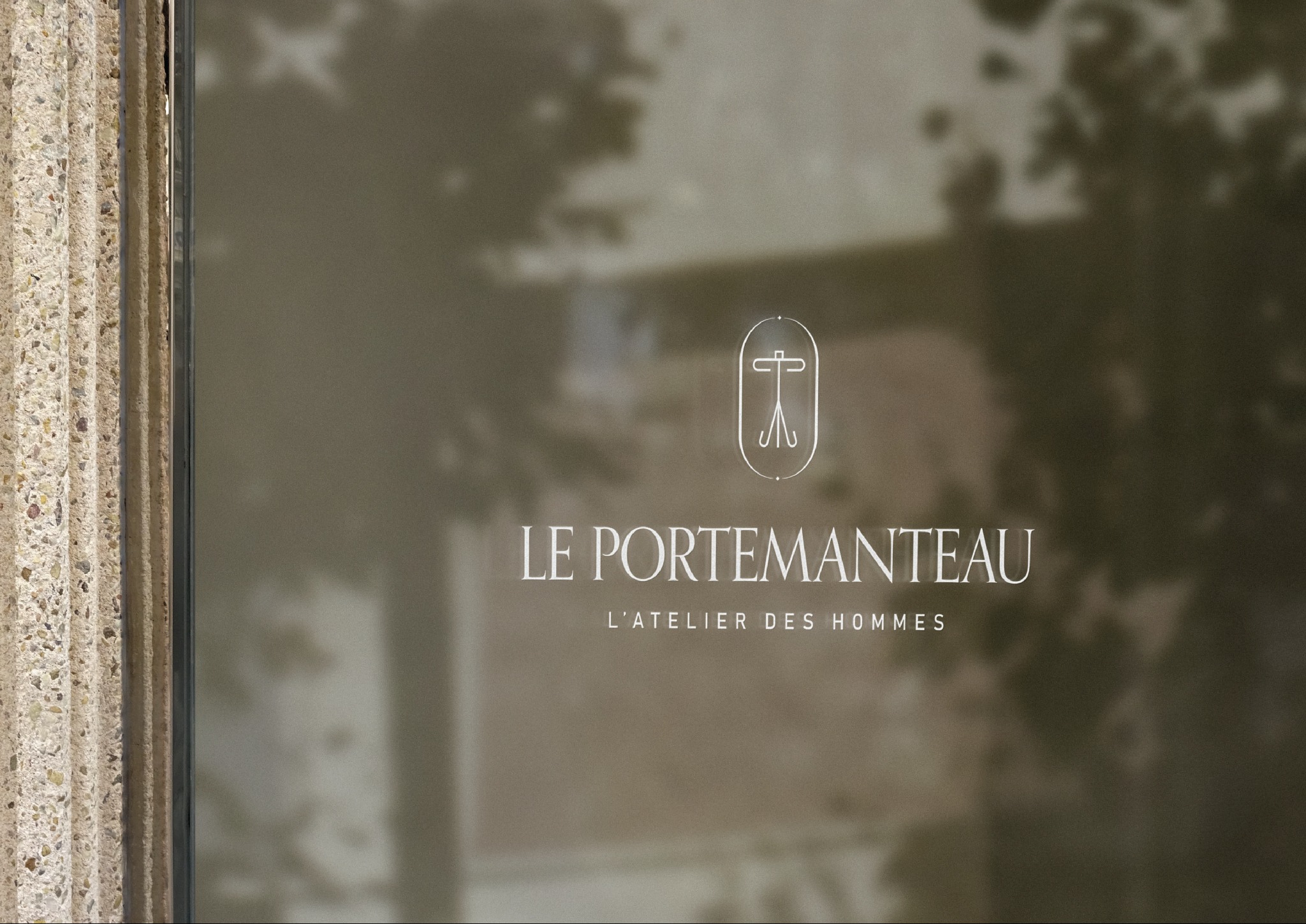 Eleganckie logo 'LE PORTEMANTEAU L'ATELIER DES HOMMES' na matowej szybie z delikatnym odbiciem otoczenia. Minimalistyczny design, stonowana kolorystyka i subtelny detal.