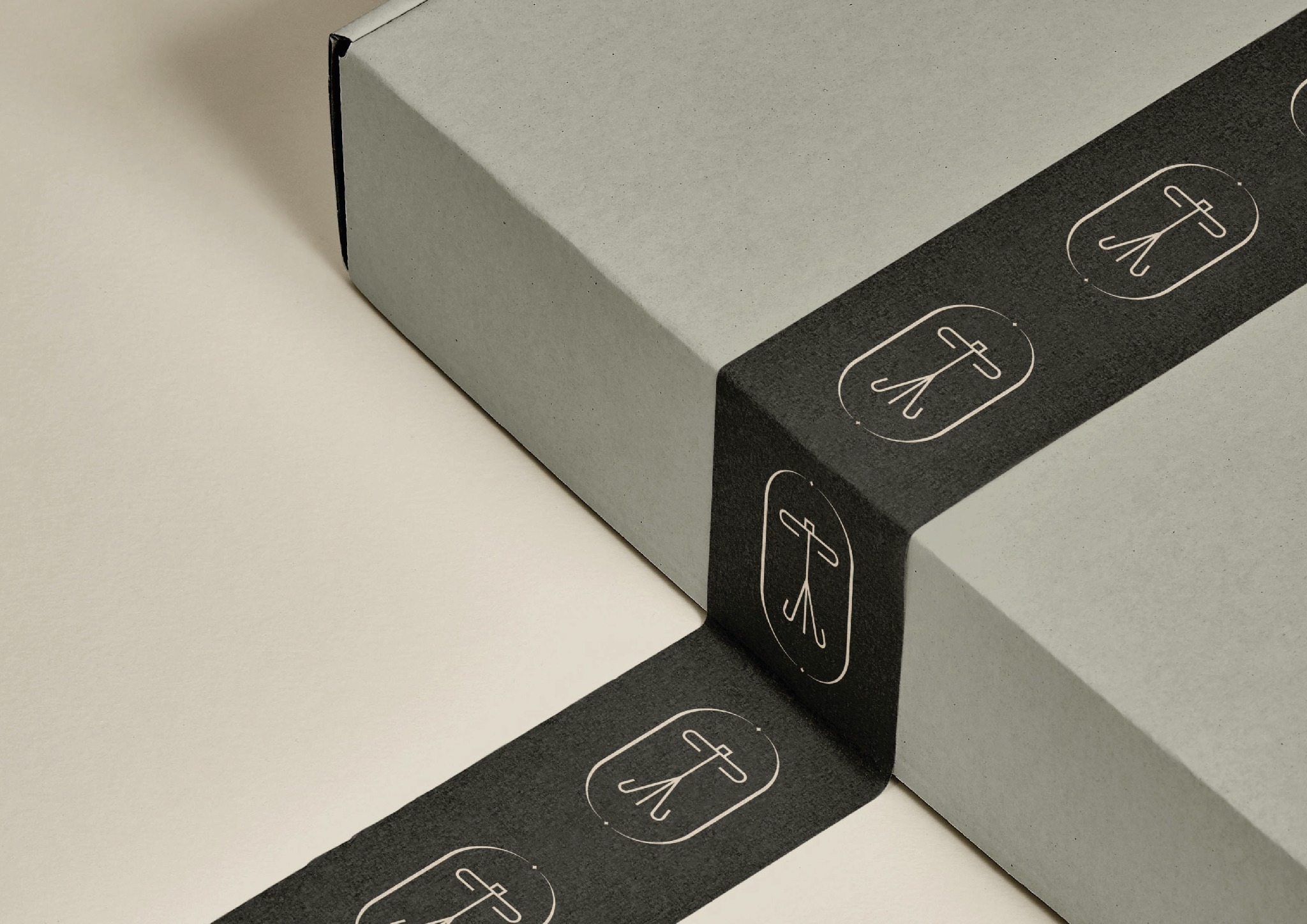 Zamknięte, szare pudełko przewiązane czarną taśmą z powtarzalnym, białym logo. Minimalistyczny design opakowania, widok z góry, neutralne tło.