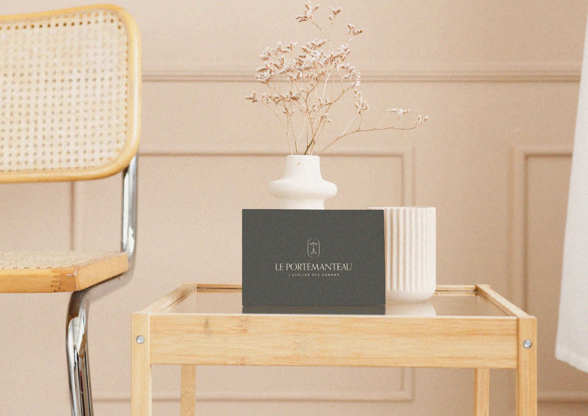 Eleganckie pudełko z logo 'Le Portemanteau' na szklanym stoliku, obok wazon z suszonymi kwiatami i krzesło w tle. Stonowana kolorystyka, minimalistyczny styl.