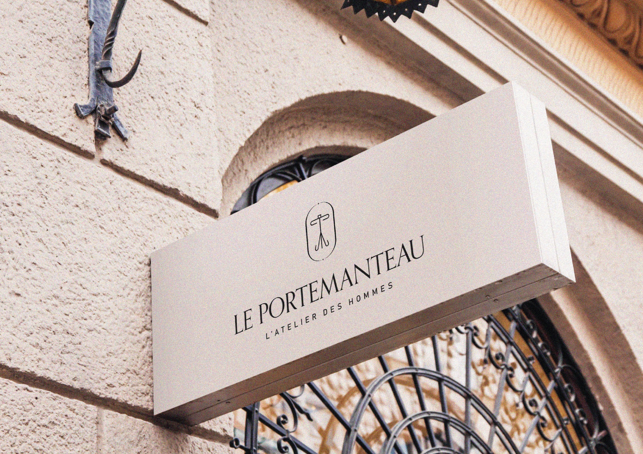 Elegancki szyld sklepu 'Le Portemanteau' z minimalistycznym logo na beżowym tle, umieszczony na teksturalnej ścianie budynku, widoczny z perspektywy żabiej.
