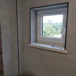 Białe okno PCV z czarną ramą osadzone w szarej, wykafelkowanej ścianie łazienki z widokiem na zieleń i budynek w oddali, nad spłuczką toalety.