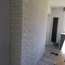 Ściana działowa wykończona białymi cegłami dekoracyjnymi, widok z boku, szara podłoga, tuba silikonu na podłodze sugeruje prace wykończeniowe.