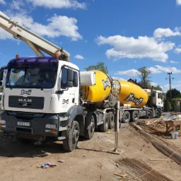 Ciężarówka MAN z żółtą gruszką do transportu betonu na placu budowy z widoczną pompą, stertą prętów zbrojeniowych i przenośną toaletą w tle, pod błękitnym niebem z chmurami.