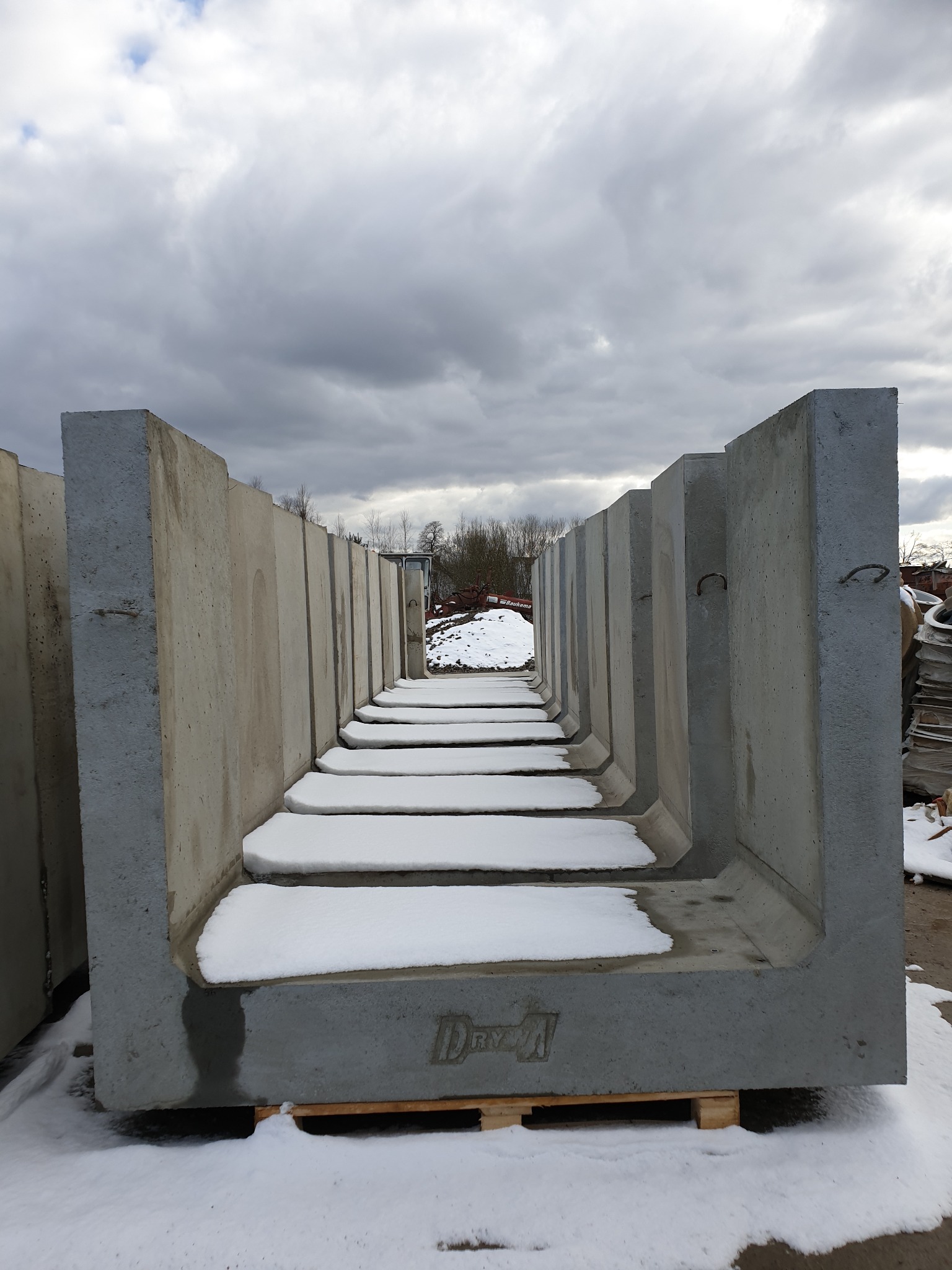 Szereg prefabrykowanych koryt betonowych z pokrywami, ułożonych na palecie, częściowo pokrytych śniegiem, na tle pochmurnego nieba.