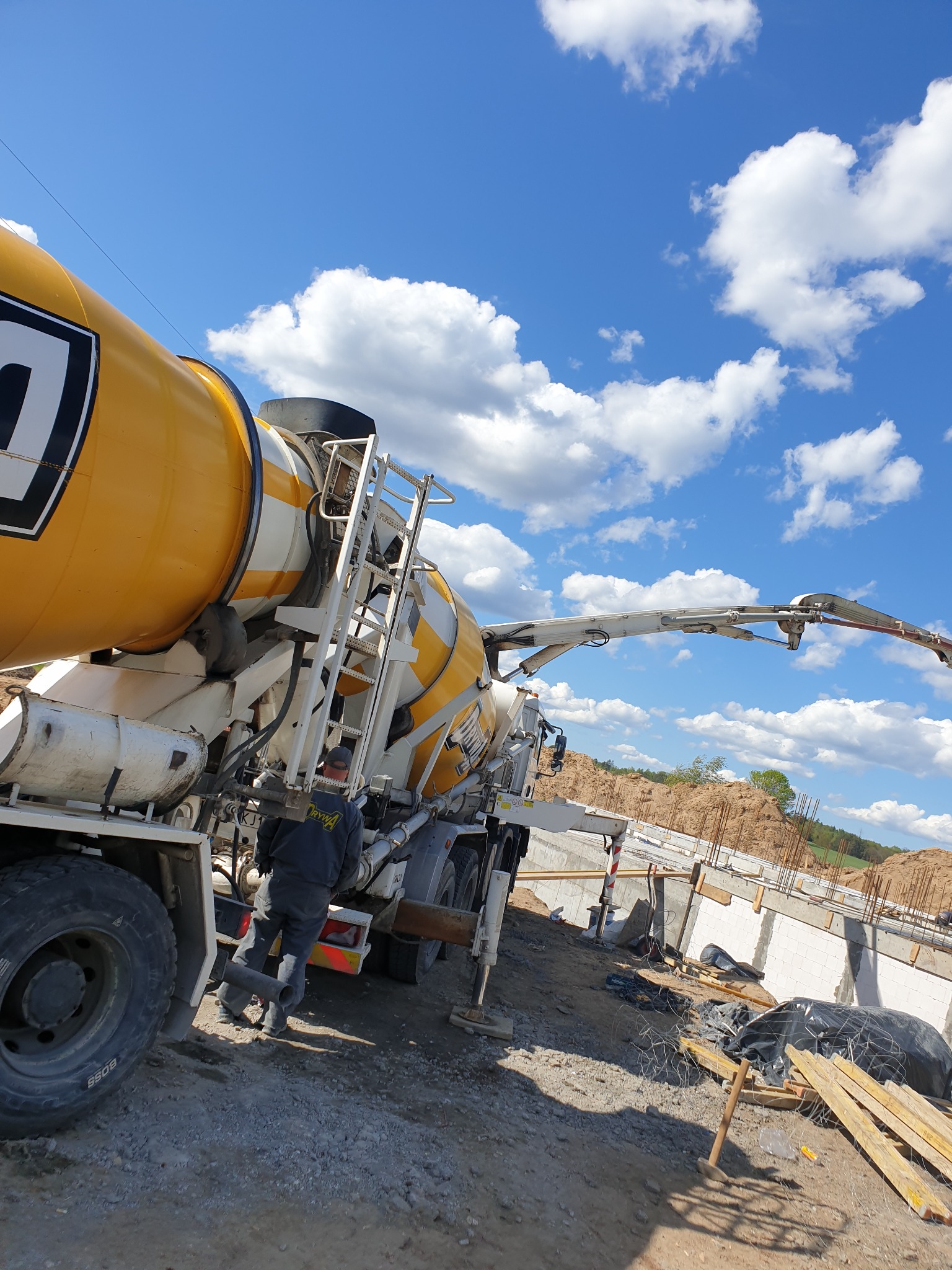 Żółto-biała betonomieszarka z wysięgnikiem na placu budowy, widoczne fundamenty i stalowe pręty zbrojeniowe, błękitne niebo z chmurami w tle.