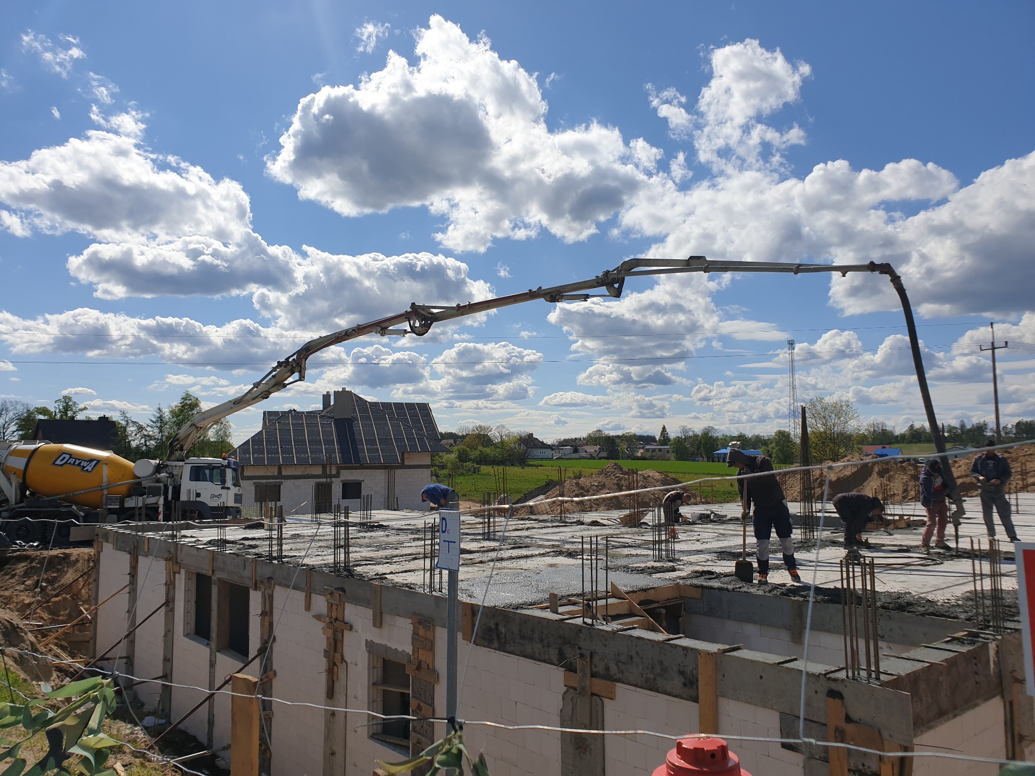 Wylewanie fundamentów domu jednorodzinnego z użyciem pompy do betonu, widoczny żółty betonomieszarka, zbrojenie i pracujący robotnicy.