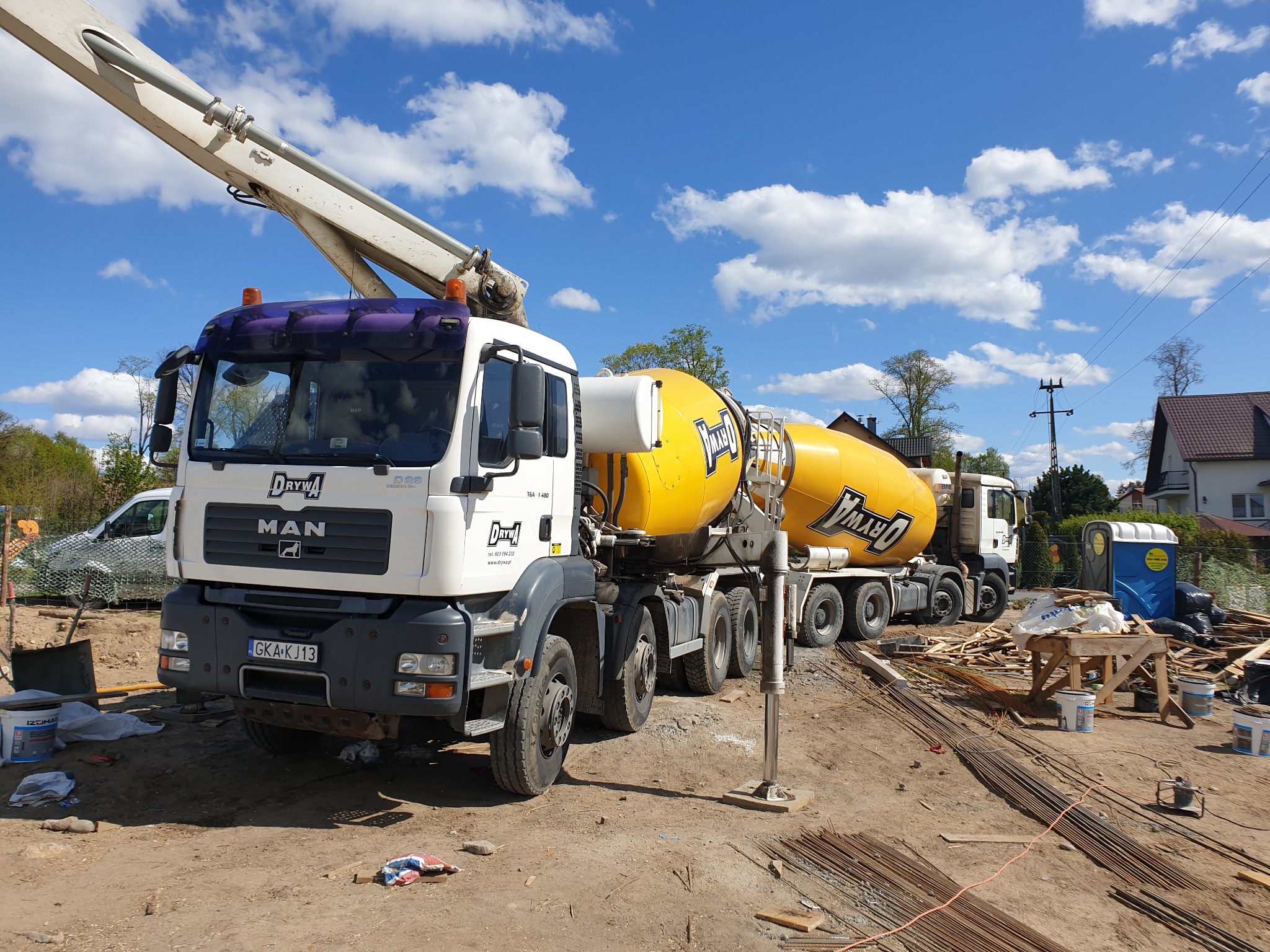 Ciężarówka MAN z żółtą gruszką do transportu betonu na placu budowy z widoczną pompą, stertą prętów zbrojeniowych i przenośną toaletą w tle, pod błękitnym niebem z chmurami.