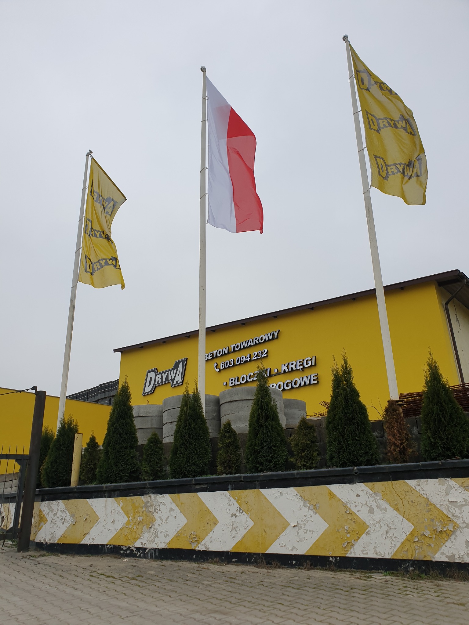 Żółty budynek firmy z logo, flagami firmowymi i polską flagą, przed budynkiem ułożone betonowe kręgi, w tle widoczna stal zbrojeniowa.