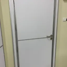 Zamknięte białe skrzydło w srebrnej ościeżnicy z poziomym panelem, widoczne zawiasy i klamka na tle beżowej ściany i szarej podłogi.