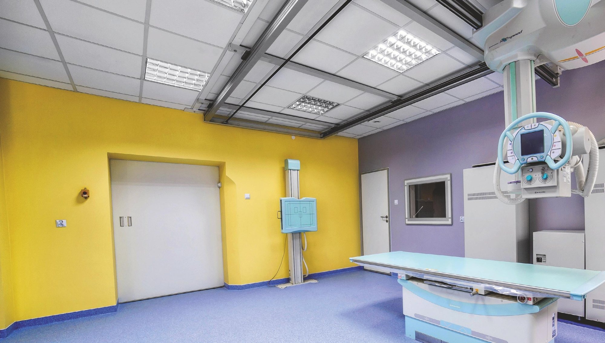 Wnętrze gabinetu rentgenowskiego z aparaturą medyczną, ścianą pomalowaną na żółto i białą przesuwną osłoną.