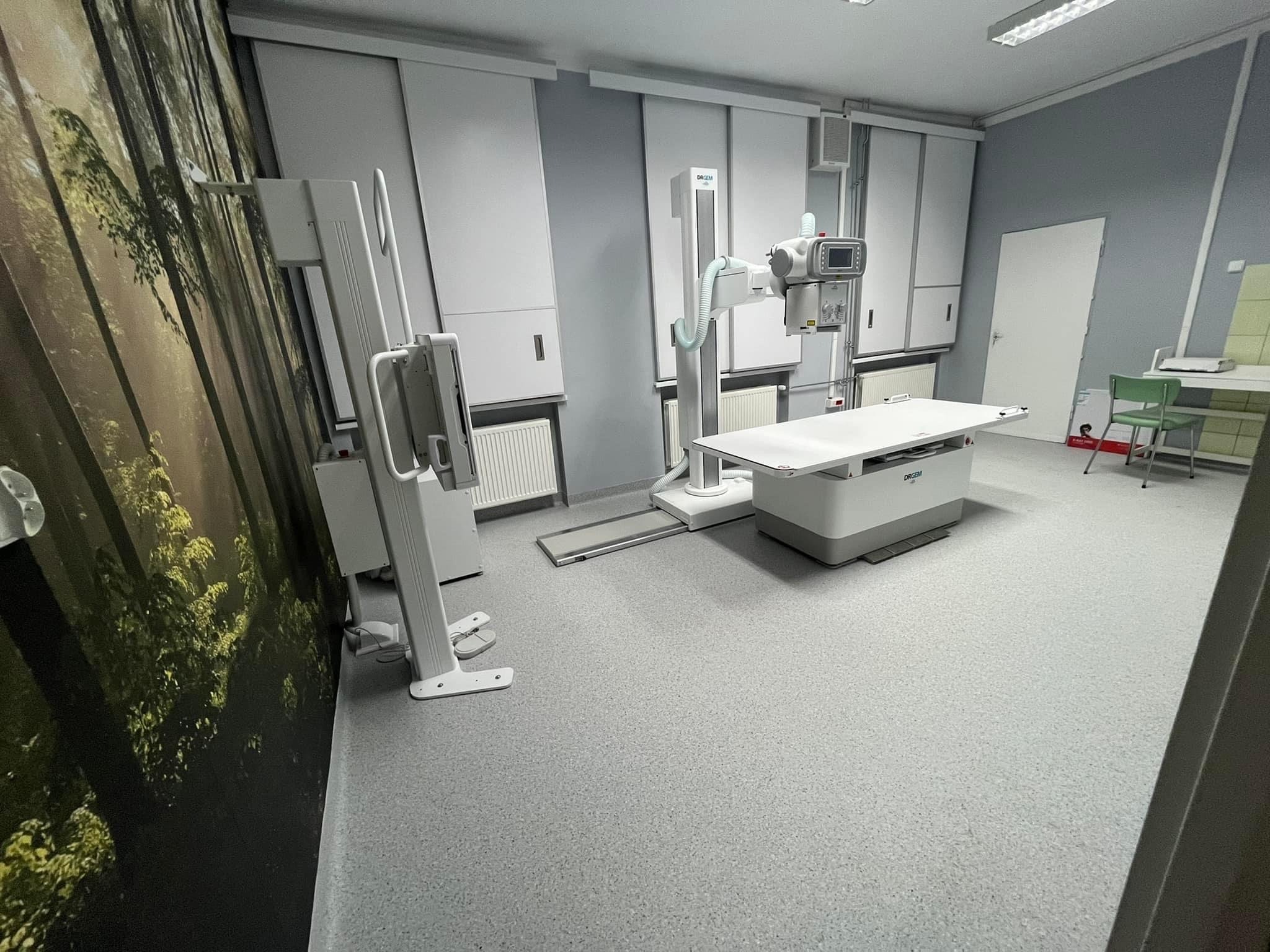 Wnętrze gabinetu z aparaturą RTG; na ścianie fototapeta z motywem leśnym, białe szafki i grzejniki, szare ściany i podłoga.
