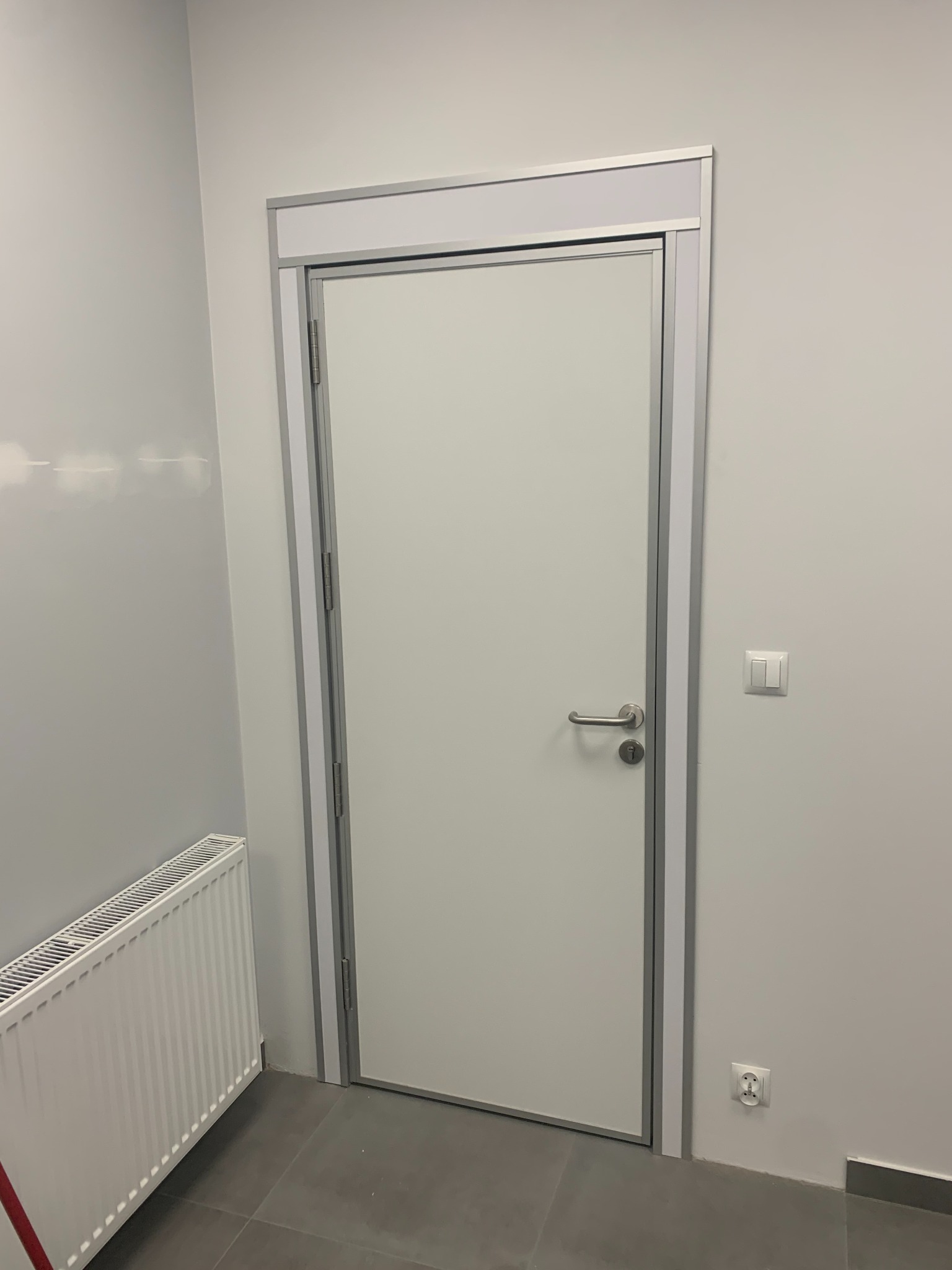 Białe skrzydło z srebrną ościeżnicą w minimalistycznym wnętrzu, widoczny grzejnik i włącznik światła.