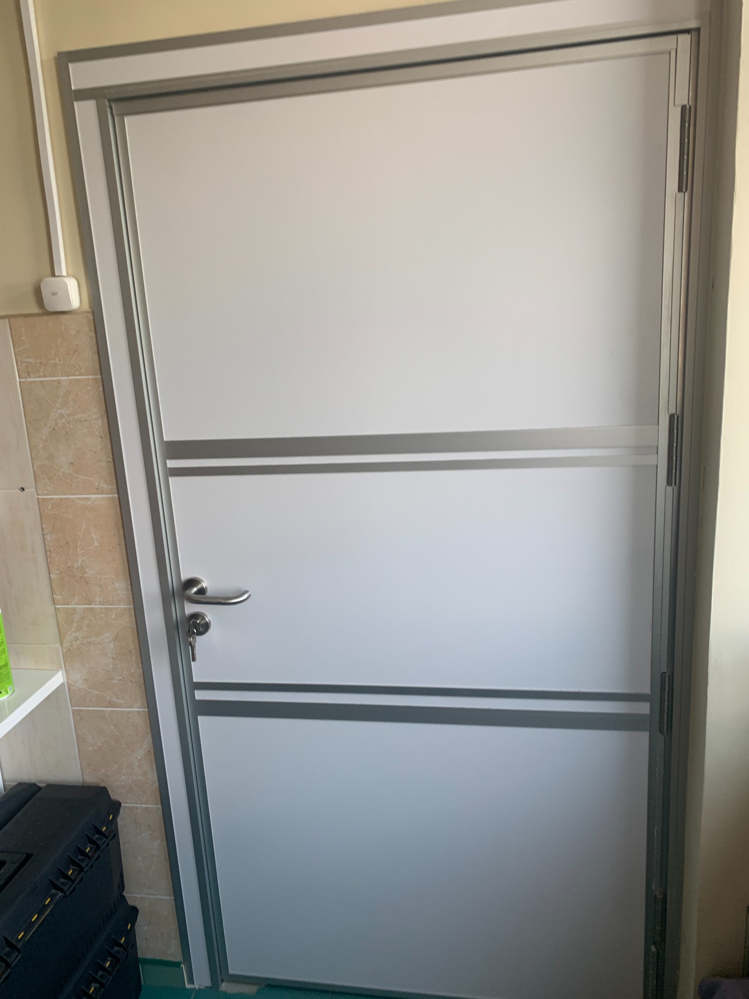 Białe skrzydło z szarymi poziomymi pasami i srebrną ramą, widoczny zamek z kluczem, fragment ściany z płytkami i czarna skrzynka narzędziowa w dolnym lewym rogu.