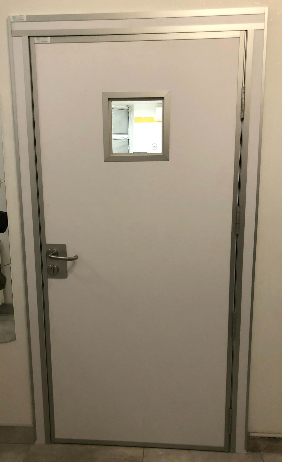 Białe drzwi wewnętrzne z małym kwadratowym okienkiem, obramowane szarą metalową ramą, widoczny srebrny uchwyt i zawiasy.