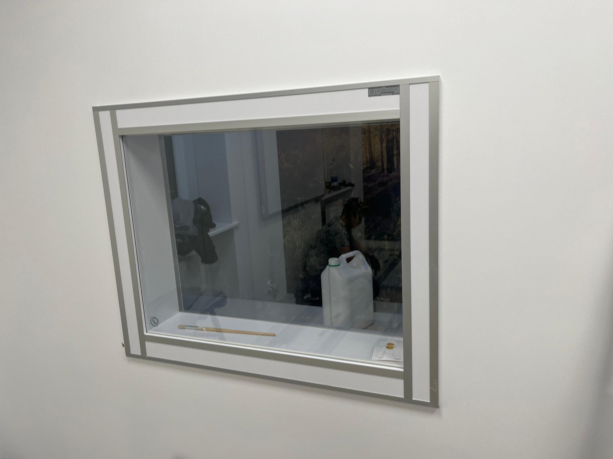 Przeszklone okienko w ścianie z ramą w kolorze szarym, za szybą widoczne przedmioty: kanister, pędzel i inne.
