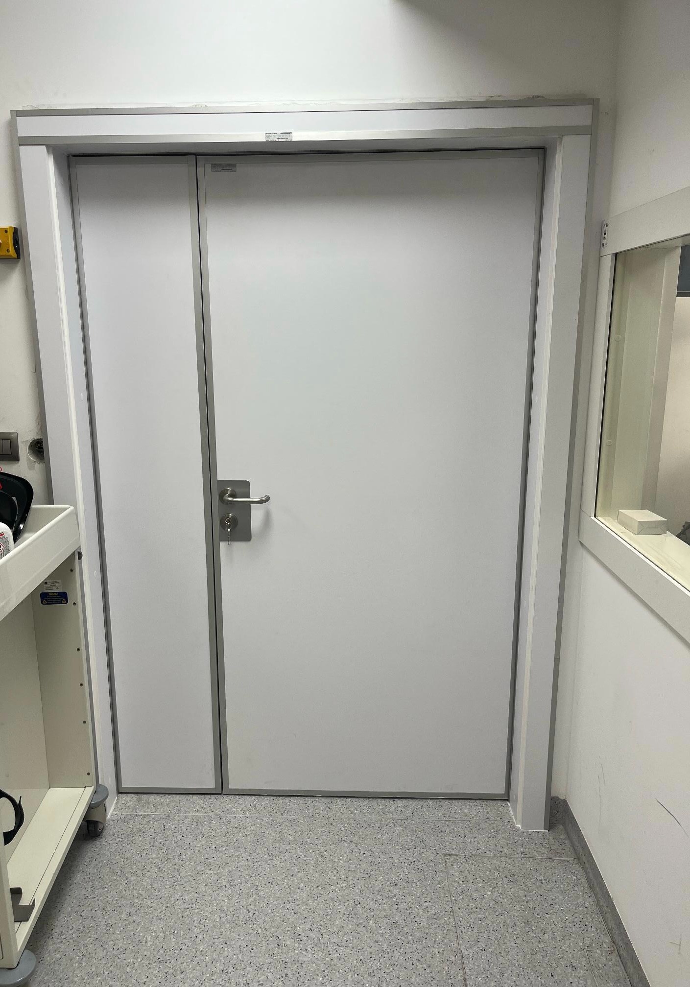 Zamknięte, białe skrzydło z szarą ramą i klamką, widziane z bliska na tle szarej podłogi i białych ścian, z fragmentem wózka medycznego po lewej stronie.