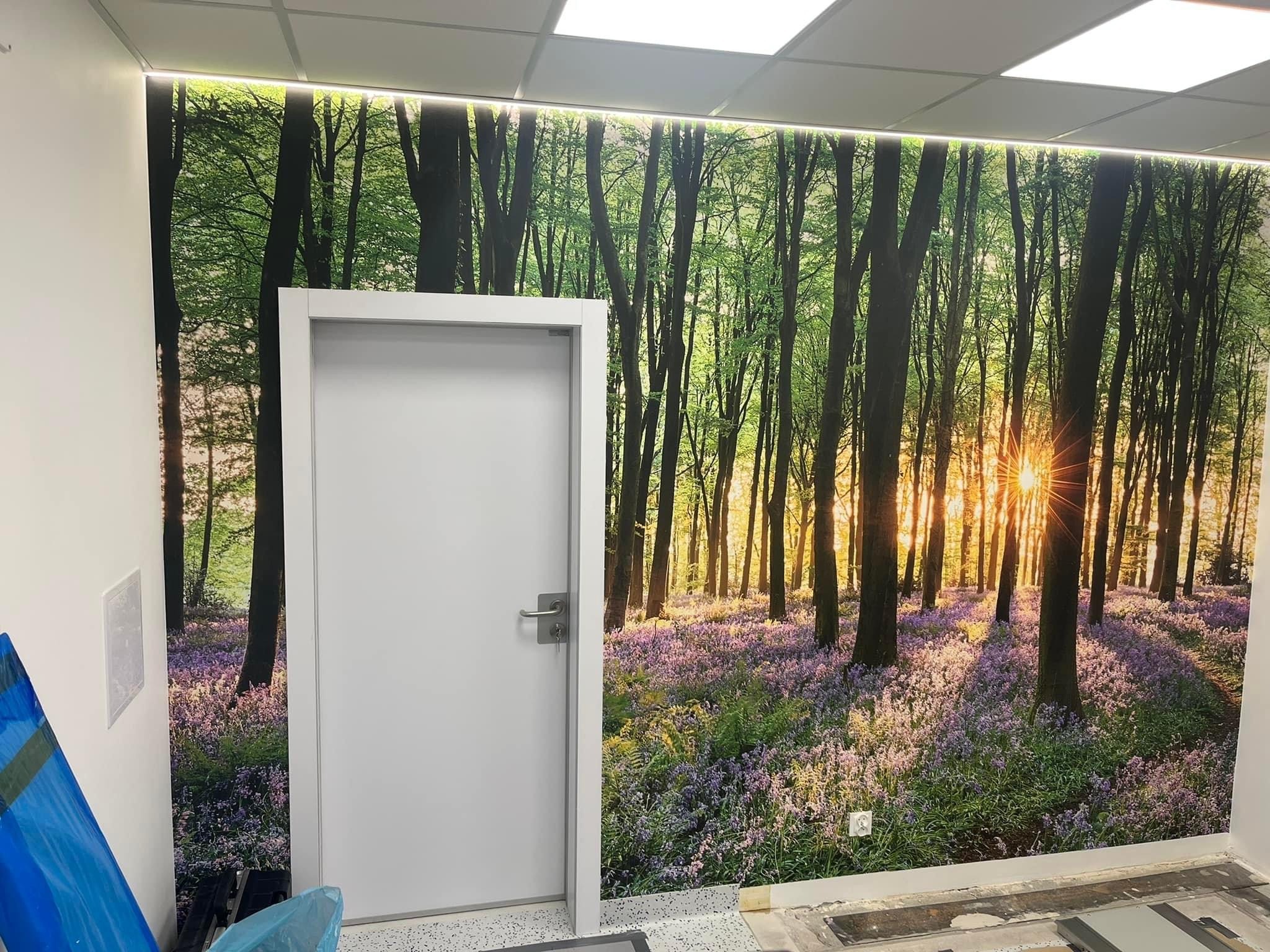 Białe skrzydło z klamką na tle fototapety z motywem leśnym i promieniami słońca, widoczne niedokończone wykończenie podłogi.