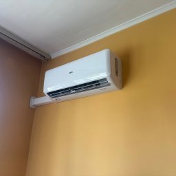 ELECTIVUS - Biała jednostka wewnętrzna klimatyzatora ściennego marki ANDE, zamontowana wysoko na jasnopomarańczowej ścianie, z widoczną białą maskownicą instalacji.