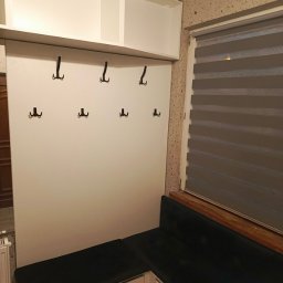 MEBLEGO SPÓŁKA Z OGRANICZONĄ ODPOWIEDZIALNOŚCIĄ - Biała zabudowa przedpokoju z szafkami wiszącymi, panelem z haczykami i siedziskiem z szufladami, uzupełniona o ciemnozieloną tapicerkę.