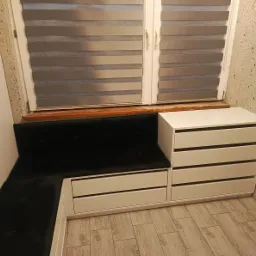 Biała zabudowa okienna z szufladami i tapicerowanym siedziskiem w kształcie litery L, z widocznymi haczykami na ścianie i żaluzjami w oknie.