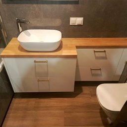 MEBLEGO - Nowoczesna łazienka z białą szafką pod umywalką z drewnianym blatem, umywalką nablatową, złotymi uchwytami, szarą płytką na ścianie i drewnopodobną podłogą.