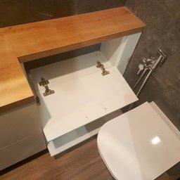 MEBLEGO SPÓŁKA Z OGRANICZONĄ ODPOWIEDZIALNOŚCIĄ - Biała szafka pod umywalkę z drewnianym blatem, otwarte drzwiczki ukazują zawiasy, obok widoczny bidet w łazience z marmurową ścianą.