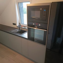MEBLEGO SPÓŁKA Z OGRANICZONĄ ODPOWIEDZIALNOŚCIĄ - Nowoczesna kuchnia w szarościach z ciemnym blatem, zintegrowana zabudowa z piekarnikiem i mikrofalówką Amica oraz czarną lodówką Samsung, widok na okno dachowe.