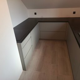 MEBLEGO SPÓŁKA Z OGRANICZONĄ ODPOWIEDZIALNOŚCIĄ - Nowoczesna kuchnia w odcieniach szarości z ciemnym blatem, minimalistyczne fronty szafek bez uchwytów, drewnopodobna podłoga, widok z perspektywy narożnika.